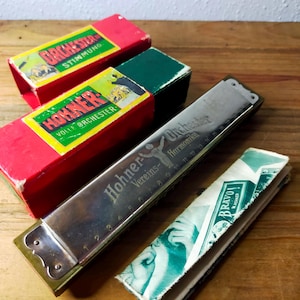 May include: A Hohner Vereins-Orchester Harmonika harmonica with a metal body, accompanied by two red and green cardboard boxes labelled Orchester Stimmung and Hohner Volle Orchester, and a green and white Bravo M. Hohner paper sleeve.