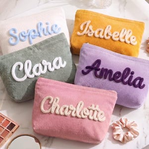 Puede incluir: Cinco estuches rectangulares de pana en color crema, amarillo mostaza, verde salvia, lavanda y rosa. Cada estuche presenta un nombre bordado en relieve: Sophie en azul claro, Isabelle en crema, Clara en blanco, Amelia en morado oscuro y Charlotte en crema.