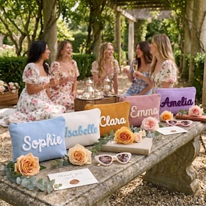 Puede incluir: Cuatro estuches rectangulares de pana en azul claro, blanco, amarillo mostaza y lavanda, cada uno con un nombre bordado en un color contrastante: Sophie, Isabelle, Clara, Emma y Amelia. Los estuches están dispuestos sobre una superficie de piedra.