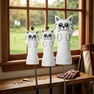 May include: Three white golf club head covers shaped like cartoon cats with gray stripes, large eyes, and small ears. Each cover features a black top knob. The covers are displayed upright on thin metal shafts, with golf tees and a glove nearby.