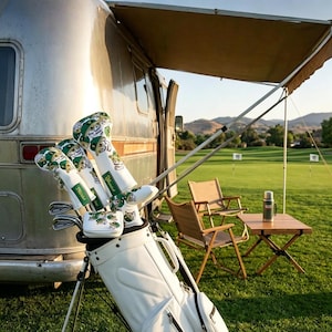 May include: A white golf bag containing several golf clubs with white headcovers featuring a green and yellow graphic pattern and the text "Ditched". The bag is positioned on a grassy field next to a silver trailer with an extended awning.