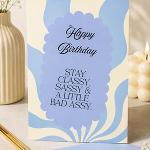 Könnte beinhalten: Eine rechteckige Grußkarte mit cremefarbenem Hintergrund und hellblauen Wellenstreifen. Zentriert ist ein hellblaues, gewelltes Oval mit dem schwarzen Text: Happy Birthday, STAY CLASSY, SASSY & A LITTLE BAD ASSY. Ein hellblauer Umschlag liegt darunter.