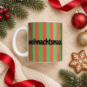 Könnte beinhalten: Eine weiße Keramiktasse mit einem vertikalen Streifenmuster in abwechselndem Grün und Rot. Der Text "weihnachtsmaus." ist in einer fetten, schwarzen, abgerundeten serifenlosen Schrift in der Mitte der Tasse aufgedruckt.