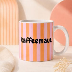 Könnte beinhalten: Ein Keramikbecher mit vertikalen orangefarbenen und hellrosa Streifen. Das Wort kaffeemaus. ist in schwarzen, kleingeschriebenen, abgerundeten serifenlosen Buchstaben in der Mitte des Bechers aufgedruckt. Henkel und Innenseite sind weiß.