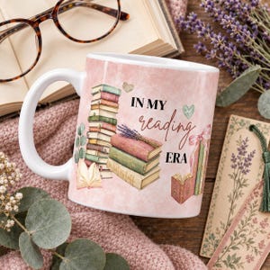 Könnte beinhalten: Ein weißer Keramikbecher mit einem zartrosa Aquarellhintergrund, bedruckt mit einem Bücherstapel und dem Text "IN MY reading ERA" in schwarzer Druck- und Schreibschrift. Das Design zeigt zudem offene Bücher, einen Lavendelzweig und kleine Herzen.