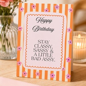 Könnte beinhalten: Eine rechteckige Grußkarte mit vertikalen orangefarbenen und weißen Streifen sowie kleinen rosa Blumen. In der Mitte befindet sich ein hellrosa Feld mit einem gewellten orangefarbenen Rand und dem Text: Happy Birthday STAY CLASSY, SASSY & A LITTLE BAD ASSY.