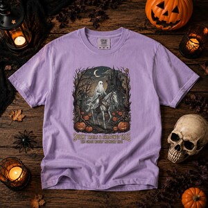 May include: A light purple short-sleeve t-shirt featuring a graphic of a ghost riding a horse through a forest with a castle, pumpkins, and a crescent moon. The text reads: SPOOKY TRAILS & SHADOWED TALES THE GHOST RIDERS MIDNIGHT RIDE.