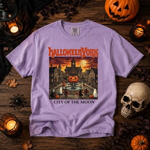 May include: A light purple short-sleeve t-shirt featuring a graphic print of a spooky town square with a large jack-o'-lantern on a fountain, ghosts, and vampires. The text reads HALLOWEEN YORK and CITY OF THE MOON in orange and white lettering.