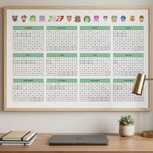 Peut inclure: Un calendrier mural 2027 dans un cadre en bois clair présentant douze grilles mensuelles avec des en-têtes verts. La partie supérieure affiche l'année 2027 en texte coloré, entourée d'une série de petites icônes de têtes illustrées.