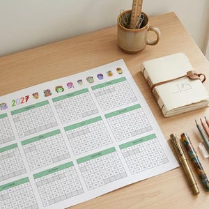 Peut inclure: Un calendrier mural 2027 présentant douze grilles mensuelles avec des en-têtes verts et de petites illustrations de personnages, accompagné d'un carnet beige avec une lanière en cuir, d'une tasse en céramique et de crayons de couleur.