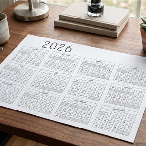 Peut inclure: Un calendrier annuel 2026 sur papier blanc présentant douze grilles mensuelles disposées en rangées. Chaque mois affiche les jours de la semaine dans un format de grille noire sur fond blanc. Le calendrier est posé sur une surface en bois.