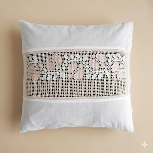May include: A square white throw pillow featuring a central horizontal band of beige and white lace. The lace panel displays a repeating floral pattern with intricate grid-like detailing and vertical lines, set against a plain white fabric background.