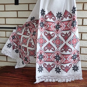 May include: A white rectangular textile runner featuring intricate cross-stitch embroidery in shades of dark gray and muted red. The pattern consists of repeating geometric diamond shapes and floral motifs, finished with a delicate white lace fringe.