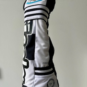 Może przedstawiać: Czarno-biały pokrowiec na kij golfowy z herbem klubu piłkarskiego Newcastle United u g&oacute;ry, pogrubionym czarnym napisem z boku, trzema poziomymi paskami oraz małą okrągłą naszywką z logo drużyny na dole z napisem AEROREADY.
