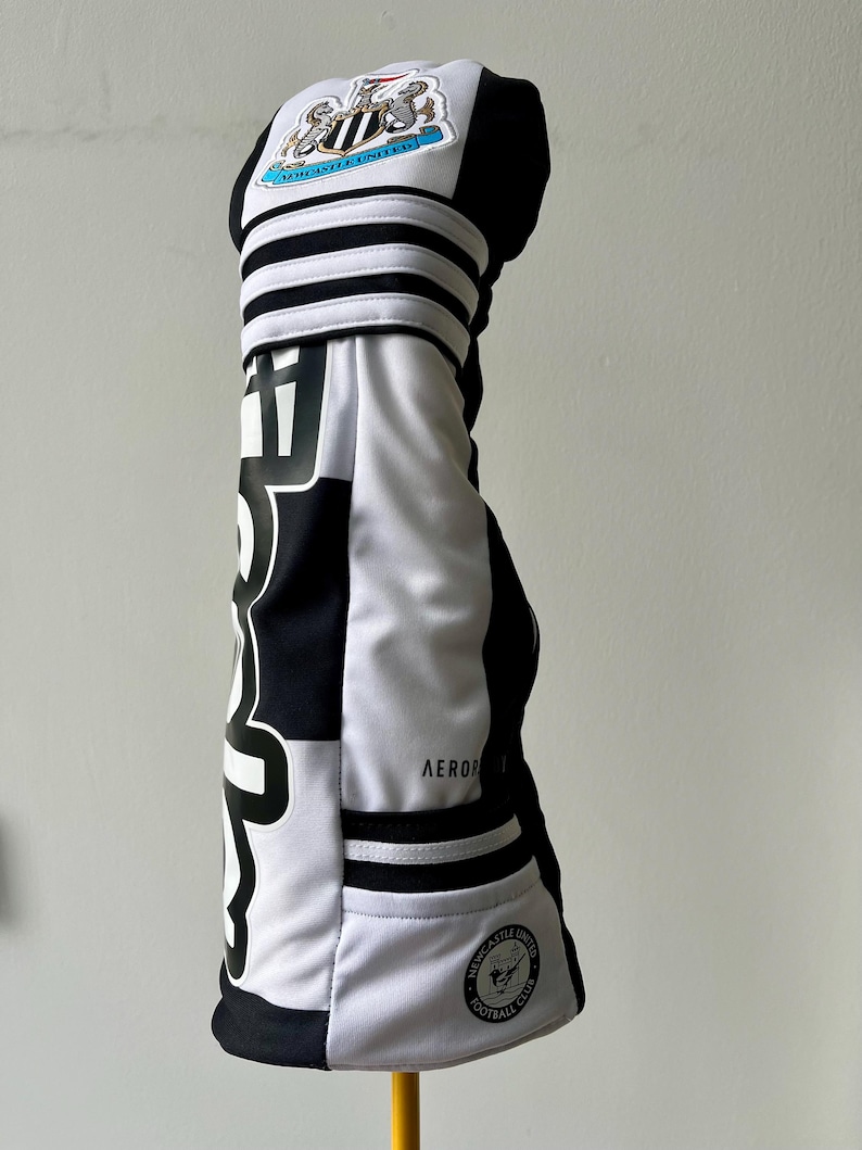Może przedstawiać: Czarno-biały pokrowiec na kij golfowy z herbem klubu piłkarskiego Newcastle United u g&oacute;ry, pogrubionym czarnym napisem z boku, trzema poziomymi paskami oraz małą okrągłą naszywką z logo drużyny na dole z napisem AEROREADY.