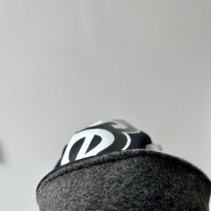 Może przedstawiać: Ciemnoszara czapka z filcowanej wełny z czarno-białym wzorem graficznym na koronie. Tkanina ma teksturowany, puszysty wygląd, a nadrukowane logo przedstawia stylizowaną białą literę B na czarnym tle.