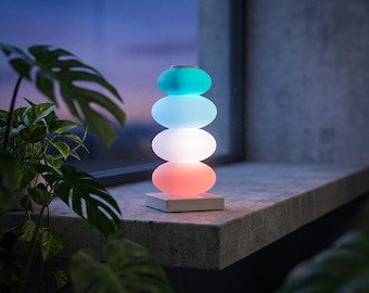 May include: A table lamp featuring four stacked, smooth, oval-shaped translucent elements that emit a gradient of light, transitioning from a vibrant teal at the top to light blue, white, and a warm coral-pink at the base, set on a square base.