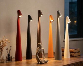 May include: A collection of six tall, slender, conical table lamps in red, black, brown, orange, gray, and white. Each lamp features a narrow base that widens slightly toward the top, topped with a small, angled light source emitting a warm glow.