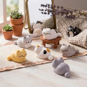 May include: A collection of small, smooth cat figurines in various colours including white, grey, yellow, and black, featuring simple facial expressions. One white cat sits on a small brown wooden stool, while others are arranged on a textured runner.