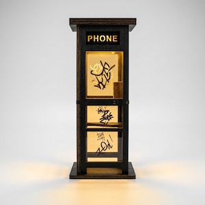 May include: A dark wood miniature telephone booth lamp with a black frame and illuminated panels. The top panel features the word PHONE in cut-out letters. The glass panels are decorated with black graffiti-style markings and emit a warm glow.