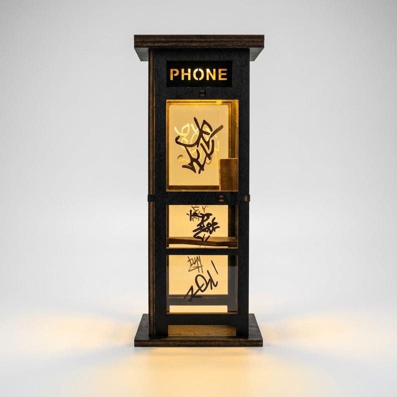 May include: A dark wood miniature telephone booth lamp with a black frame and illuminated panels. The top panel features the word PHONE in cut-out letters. The glass panels are decorated with black graffiti-style markings and emit a warm glow.