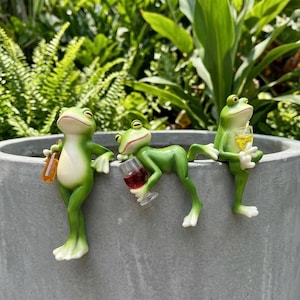 May include: Three green frog figurines perched on a grey rim. The left frog holds an amber bottle, the centre frog reclines while holding a glass of red liquid, and the right frog sits upright holding a yellow drink with a straw in a clear glass.