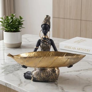 Op de afbeelding: Een decoratief beeldje van een persoon in een zwart-gouden outfit met bladpatroon, met grote oorringen en een hoofdtooi, die een grote, langwerpige, goudkleurige schaal in de vorm van een blad in de handen houdt.