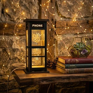 May include: A black, rectangular, lantern-style lamp shaped like a telephone booth with the word PHONE printed at the top. The lamp features clear panels with etched white line designs and emits a warm, internal glow from its base.