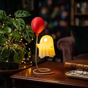 Könnte beinhalten: Eine dekorative Lampe in Form eines gelben Geistes, der einen roten Ballon hält. Der Geist hat zwei kreisförmige Augenausschnitte und ist an einem geschwungenen, hellen Metallständer befestigt, der in einem runden Fuß endet.