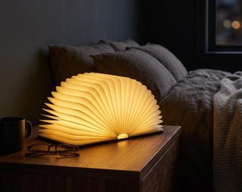 May include: A book-shaped lamp with a wooden cover and accordion-style pages that emit a warm, soft glow. The lamp is open in a semi-circular shape, resting on a wooden surface next to a black mug and a pair of dark-rimmed reading glasses.