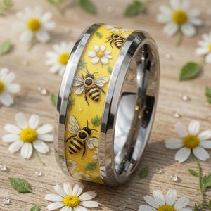 Peut inclure: Une bague en métal argenté avec une bande centrale jaune ornée d'un motif répétitif d'abeilles, de fleurs de marguerites blanches et de petites feuilles vertes. La bague présente des bords métalliques polis et un profil lisse et arrondi.