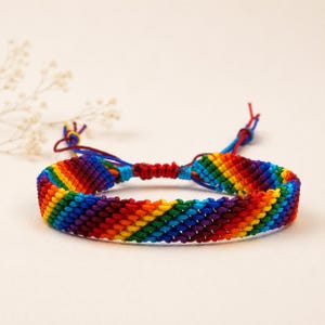 May include: A woven friendship bracelet featuring a diagonal rainbow pattern with vibrant stripes of red, orange, yellow, green, blue, and purple thread. The bracelet includes adjustable knotted cords at the back for a secure fit.