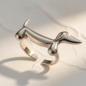 Puede incluir: Un anillo de metal plateado pulido con forma de perro salchicha hecho de globos. La figura del perro presenta una superficie lisa y reflectante con una pequeña piedra preciosa redonda y transparente engastada en la cabeza como el ojo.