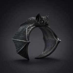 May include: A dark silver-toned ring shaped like a bat with its wings spread wide. The bat features detailed fur texture on its head and back, with intricate lines defining the structure of its wings. The ring has an open band design.