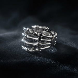 Op de afbeelding: Een zilverkleurige ring in de vorm van een skeletachtige hand die om de vinger wikkelt. Het ontwerp bevat gedetailleerde, gearticuleerde vingerkootjes en gewrichten, met een gepolijste metalen afwerking die licht reflecteert op de gebogen botstructuren.