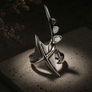 Peut inclure: Une bague en argent poli présentant un design sculptural allongé qui s'étire vers le haut. La surface métallique est lisse et réfléchissante, mettant en valeur une structure pointue avec trois protubérances arrondies en forme de feuilles.