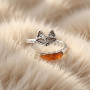 Könnte beinhalten: Ein silberfarbener offener Ring mit einem Fuchskopf-Design, strukturierten Metalldetails und einem glatten, bernsteinfarbenen, durchscheinenden Steinakzent in Form eines Fuchsschwanzes, der sich um den Finger legt.