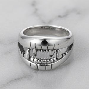 May include: A polished silver-tone ring featuring a detailed design of an open mouth with visible teeth and prominent fangs. The metal surface is smooth and reflective, showcasing the anatomical structure of the upper and lower jaw lines.