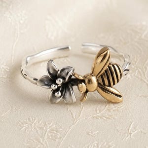 Könnte beinhalten: Ein verstellbarer offener Ring mit einer polierten silberfarbenen Blume mit sechs Blütenblättern und einer goldfarbenen Biene mit detaillierten Flügeln und Körpersegmenten. Die Ringschiene hat eine strukturierte Oberfläche.