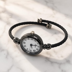 Op de afbeelding: Een horloge met een zwarte kabelarmband, een ronde kast, een witte wijzerplaat, zwarte wijzers en zwarte Romeinse cijfers. Op de wijzerplaat staat de tekst QUARTZ en de armband is afgewerkt met twee kleine zwarte metalen eindkapjes.