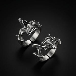 Op de afbeelding: Twee zilverkleurige metalen ringen met een gestileerd schedelontwerp. Elke ring heeft een open bandconstructie met een geborstelde afwerking, met gedetailleerde botstructuren en tanden. De ringen zijn in verschillende hoeken geplaatst.