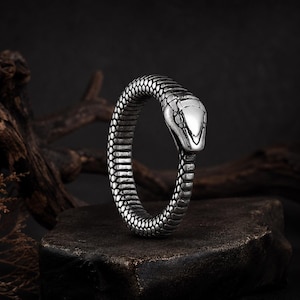 Puede incluir: Un anillo de metal plateado con forma de serpiente enroscada. La banda presenta un patrón de escamas detallado y texturizado que envuelve el dedo, con la cabeza de la serpiente formando el punto focal del diseño en metal liso y pulido.