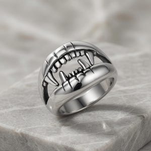 May include: A silver-toned metal ring shaped like an open mouth with prominent fangs and visible teeth. The design features a polished finish with dark recessed areas between the teeth to define the structure of the upper and lower jaw.