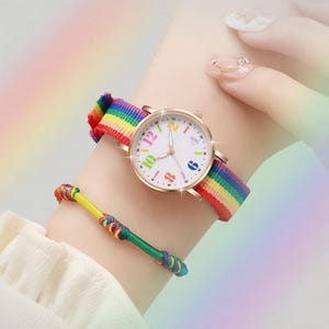Peut inclure: Une montre-bracelet avec un boîtier rond doré rose, un cadran blanc avec des chiffres multicolores et un bracelet en nylon aux rayures arc-en-ciel. Un fin bracelet en cordon vert avec des accents tissés multicolores est placé sous la montre.