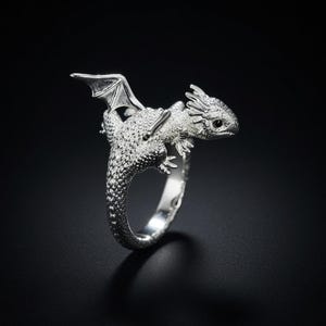 Peut inclure: Une bague en argent en forme de petit dragon avec des écailles texturées, une aile visible et une tête détaillée. Le corps du dragon forme l'anneau de la bague et sa queue se courbe pour rejoindre la tête, créant un design sculptural continu.