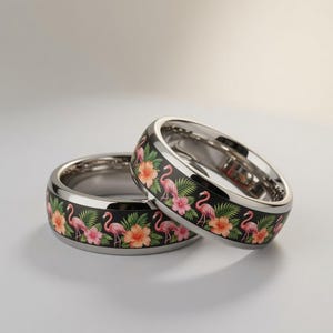 Peut inclure: Deux bagues métalliques avec une bande incrustée noire ornée d'un motif répétitif de flamants roses, de fleurs d'hibiscus orange et roses, et de feuilles de palmier tropicales vertes. Les bagues ont une finition métallique argentée polie.