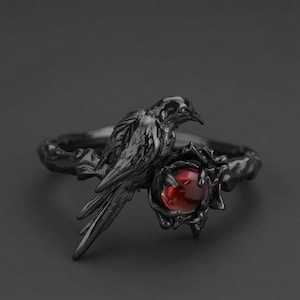 Peut inclure: Une bague en métal noir représentant un corbeau sculpté perché sur un anneau texturé. Le corbeau est placé à côté d'une petite pierre précieuse ronde rouge foncé, maintenue dans un sertissage sombre aux formes organiques rappelant des branches.