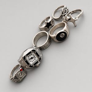 Peut inclure: Une collection de bagues argentées comprenant une bague montre avec un cadran blanc et des chiffres noirs, une bague avec une incrustation de boule huit, une bague en forme d'étoile noire, une bague avec une pierre rouge et deux anneaux texturés.