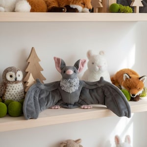 Könnte beinhalten: Ein Fledermaus-Plüschtier mit dunkelgrauen Flügeln, einer hellgrauen flauschigen Brust, rosa Innenohren und einer rosa Nase. Die Fledermaus steht auf einem Holzregal neben einer Eule, einem weißen Kaninchen, einem Fuchs und hölzernen Baumdekorationen.