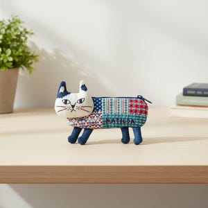 Peut inclure: Une trousse en tissu en forme de chat avec une tête blanche aux marques noires et un corps recouvert d'un motif patchwork multicolore rouge, bleu et vert. Le nom MATILDA est brodé en fil bleu foncé sur le côté du corps de la trousse.